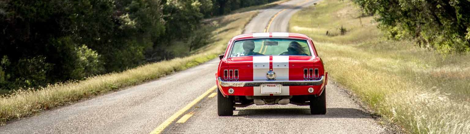 Classic Ford Musang with white stripes on a scenic road surrounded by trees